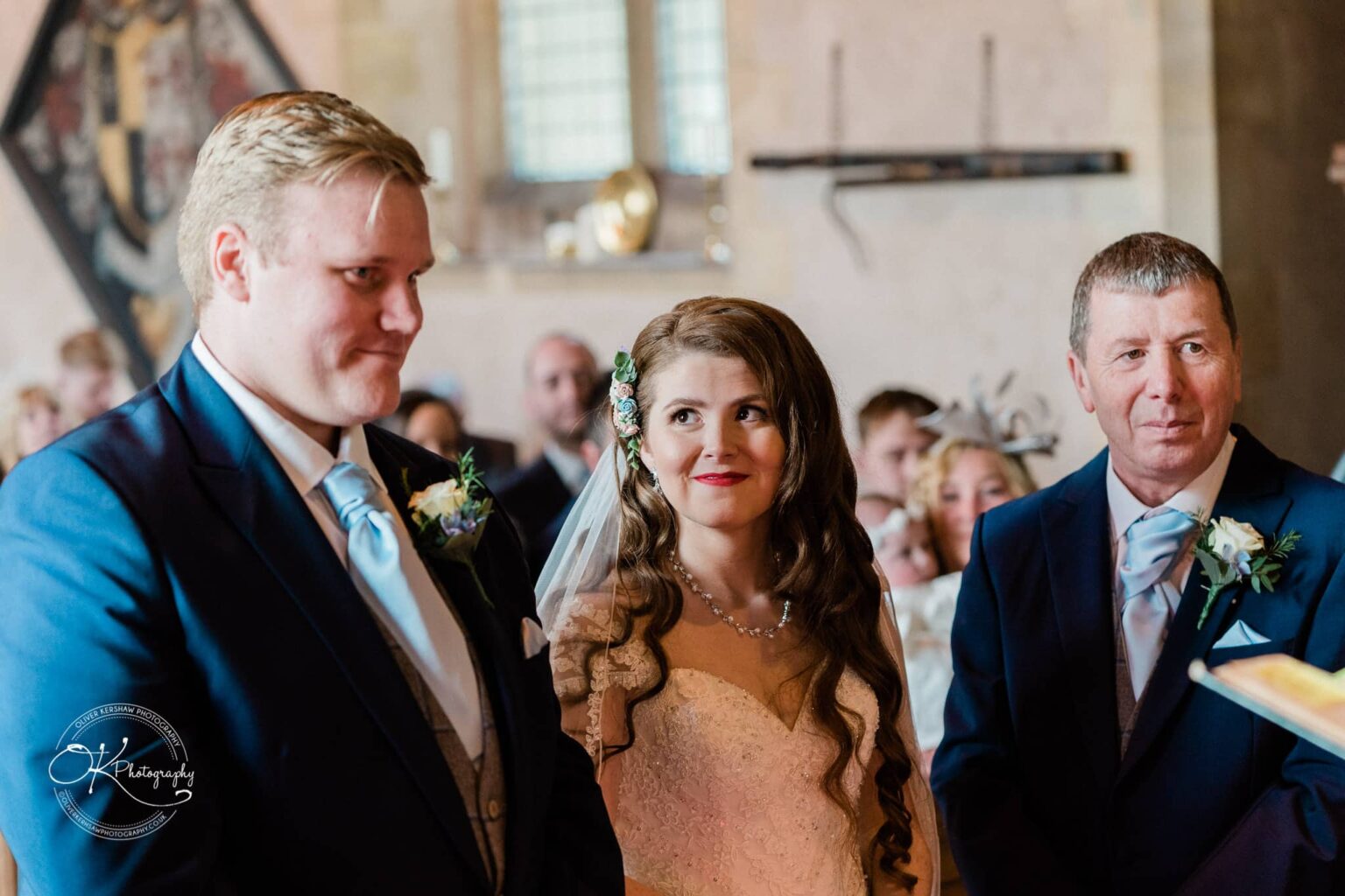 Prestwold Hall Wedding Photography A bride and groom stand together during their wedding ceremony, accompanied by an older gentleman, all dressed in formal attire with flower boutonnieres.