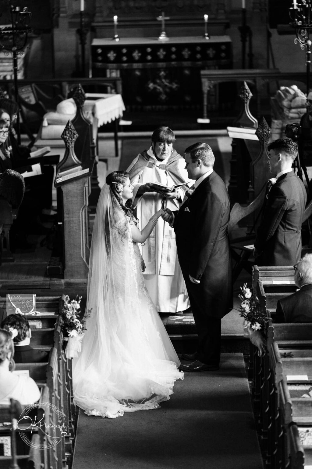 Prestwold Hall Wedding Photography A bride and groom exchanging rings in a church ceremony officiated by a priest, with guests seated in pews.
