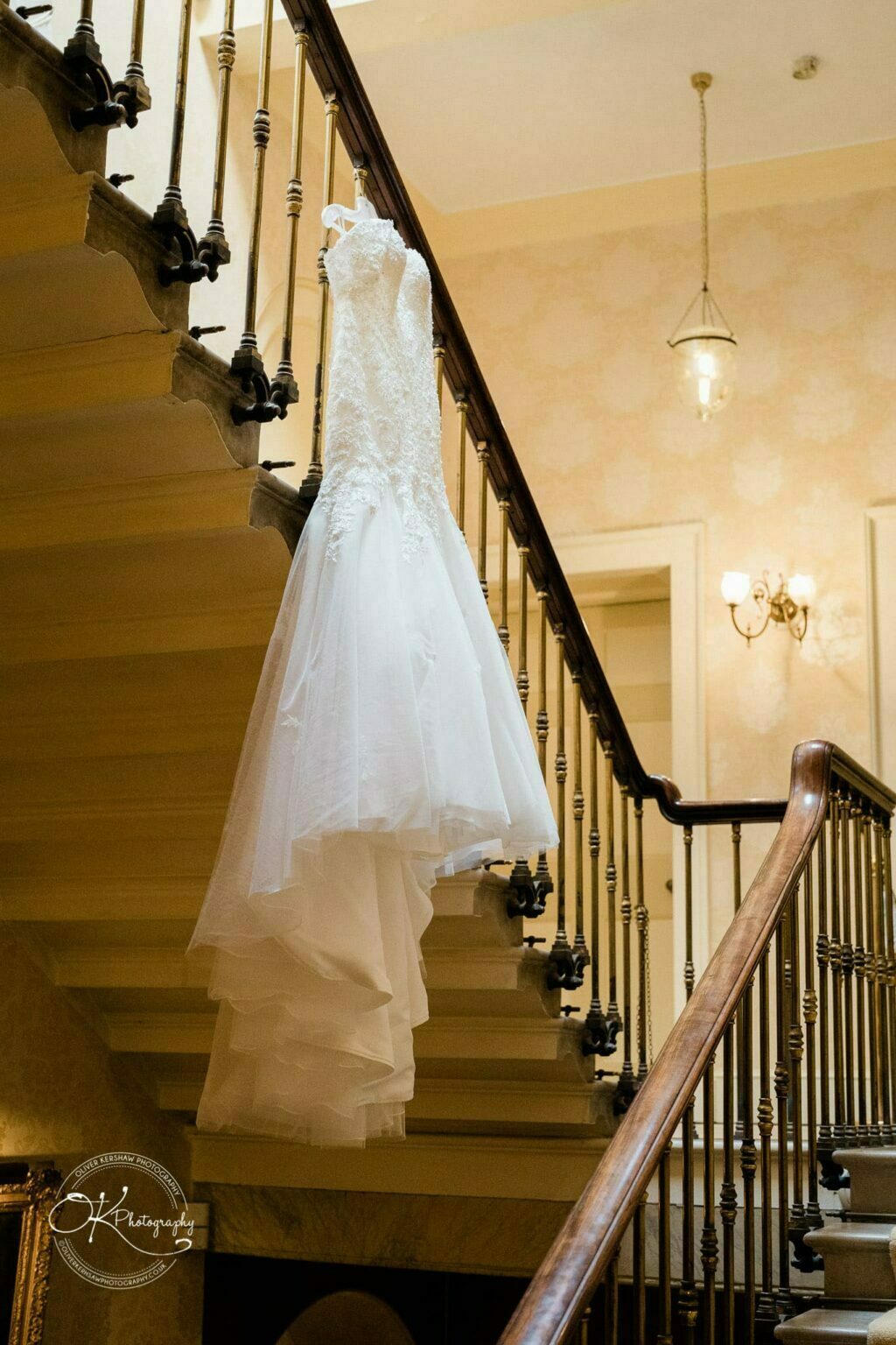 Prestwold Hall Wedding Photography A wedding dress hanging on a stair railing in a grand interior with brass balusters and ornate lighting.
