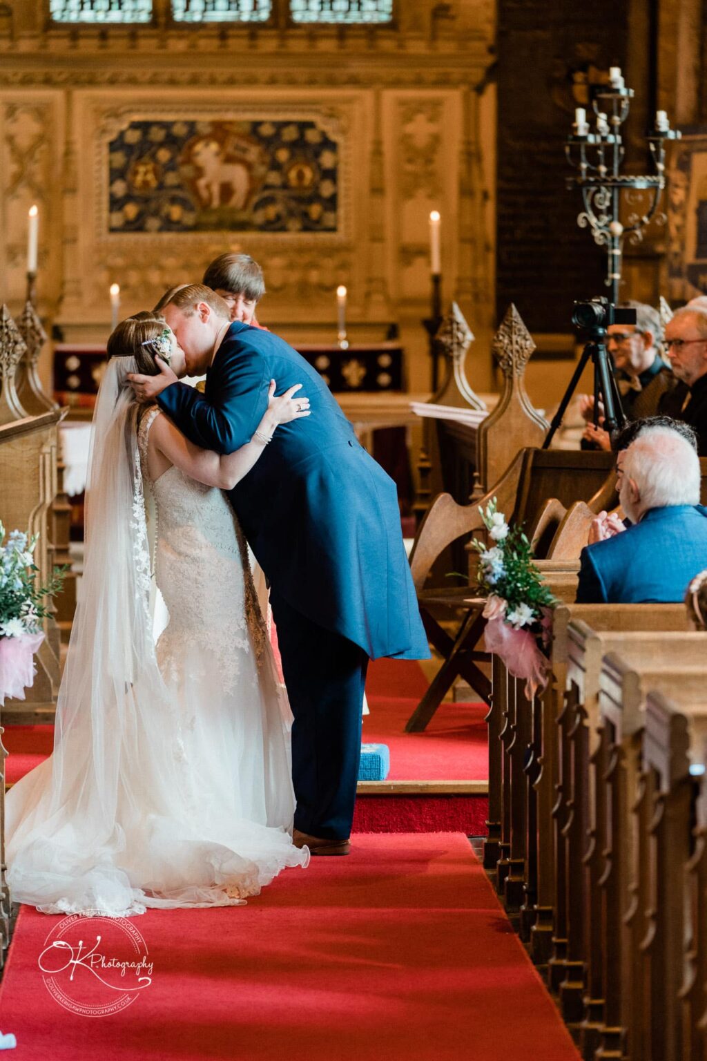 Prestwold Hall Wedding Photography A bride and groom share their first kiss during their wedding ceremony in a beautifully decorated chapel with stained glass windows and candle holders.