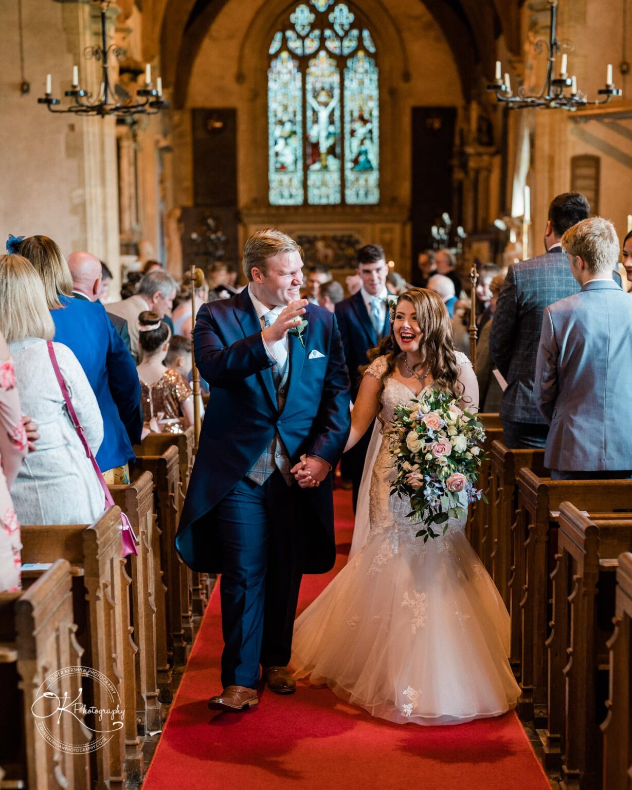 Prestwold Hall Wedding Photography A newlywed couple walking down the aisle of a church, smiling and holding hands, surrounded by guests.