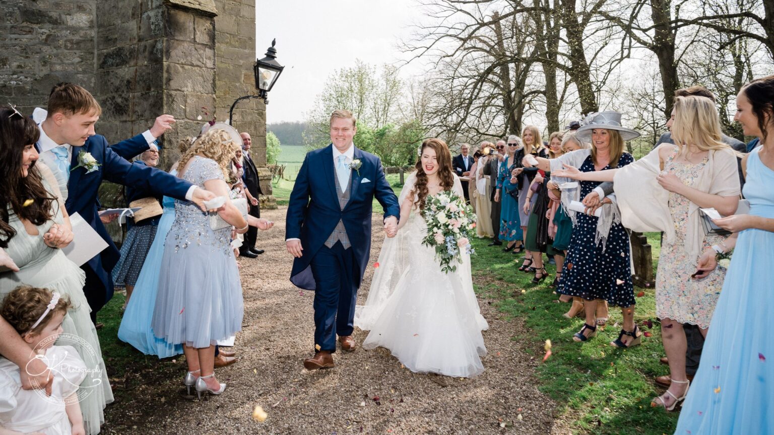 Prestwold Hall Wedding Photography A newly married couple holding hands and walking down an outdoor path, surrounded by guests throwing confetti at Prestwold Hall.