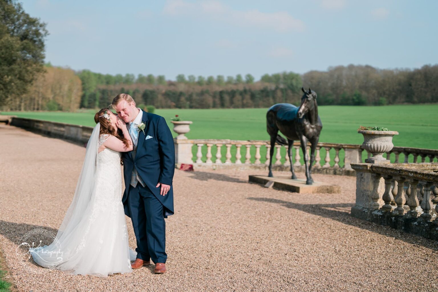 Prestwold Hall Wedding Photography A bride and groom embracing outdoors on a gravel path with a green lawn, forest, and a horse statue in the background.