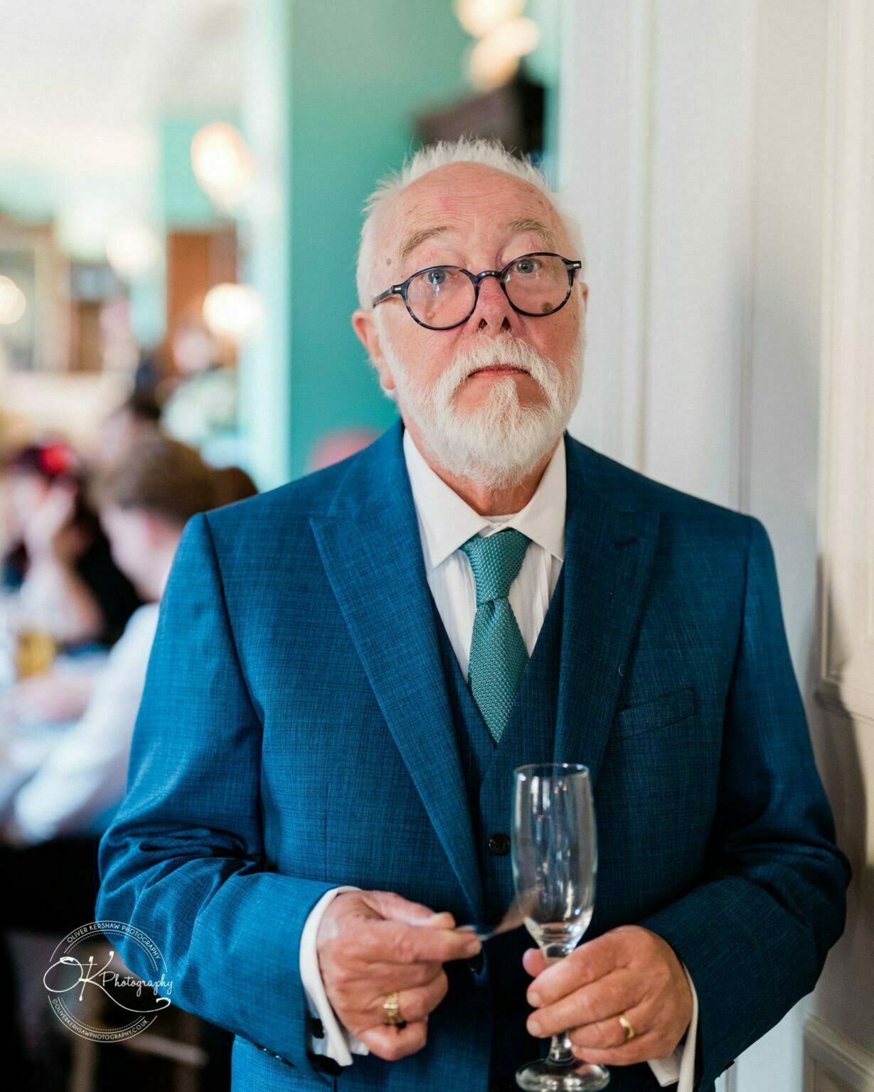 Prestwold Hall Wedding Photography Elderly man in a blue suit holding a champagne flute and a spoon inside Prestwold Hall.