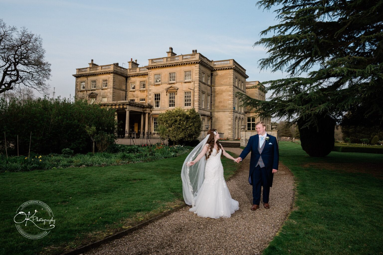 Prestwold Hall Wedding Photography A newlywed couple holding hands and walking on a path in front of a grand country house, Prestwold Hall, surrounded by lush greenery and gardens.
