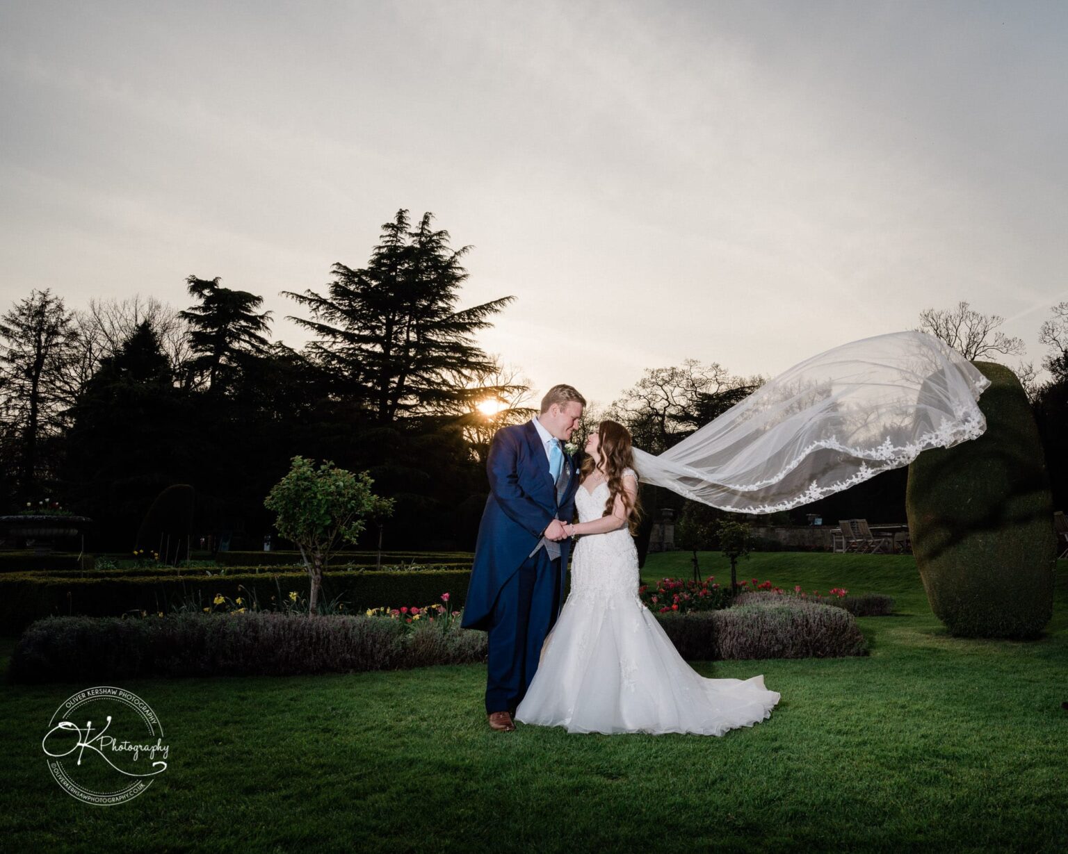 Prestwold Hall Wedding Photography A bride and groom stand together in an outdoor garden with tall trees and a sunset in the background, the bride's veil flowing in the wind.