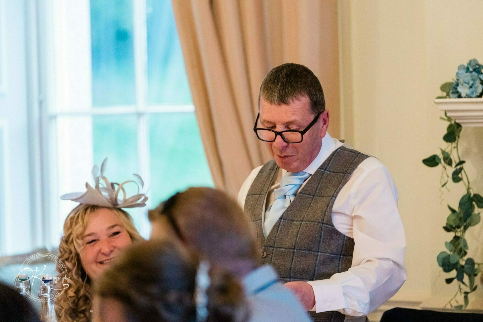 Prestwold Hall Wedding Photography A man in a plaid vest and glasses, reading from a paper at a formal event, with a woman in a fascinator looking on and smiling in a room with beige curtains and greenery.