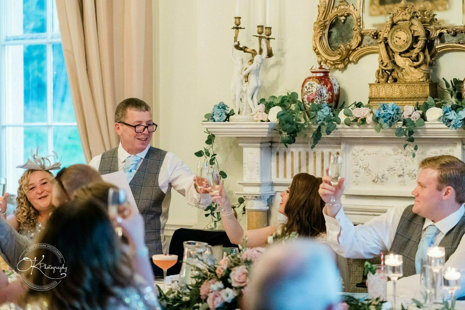 Prestwold Hall Wedding Photography People raising their glasses in a toast at a formal event, in an elegantly decorated room with floral arrangements and ornate decor.