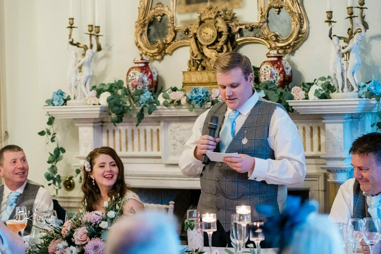 Prestwold Hall Wedding Photography A wedding speech being given in a decorated room with a fireplace and flowers, with the bride and guests seated and listening.