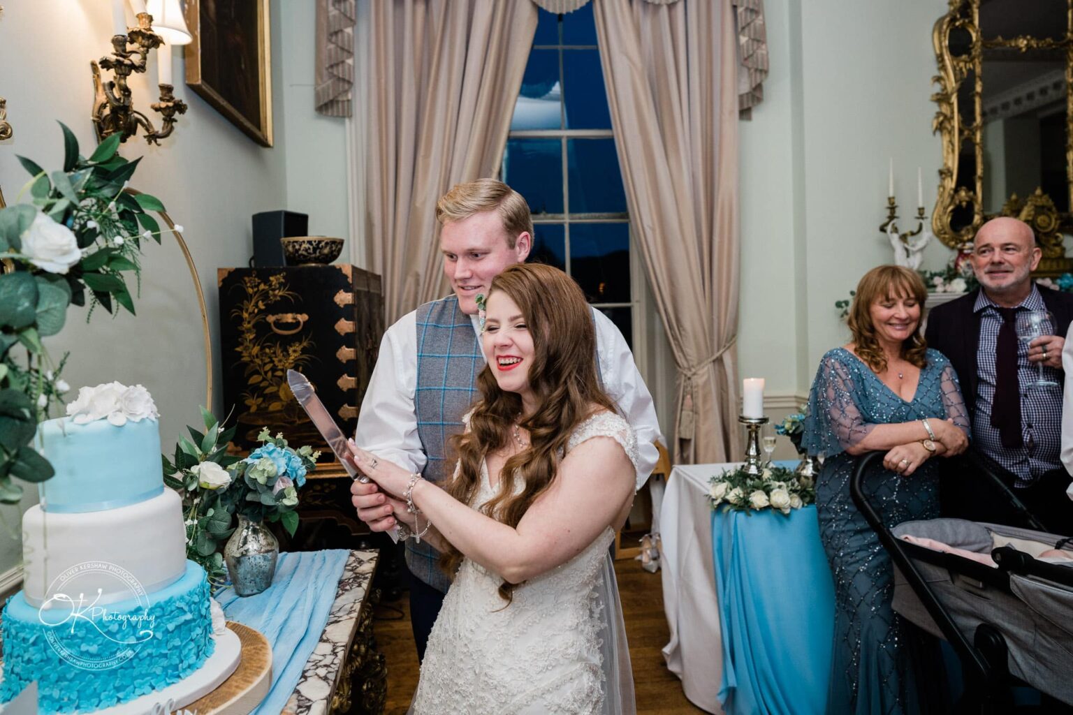 Prestwold Hall Wedding Photography A smiling bride and groom are cutting their wedding cake at Prestwold Hall, surrounded by guests in an elegantly decorated room.