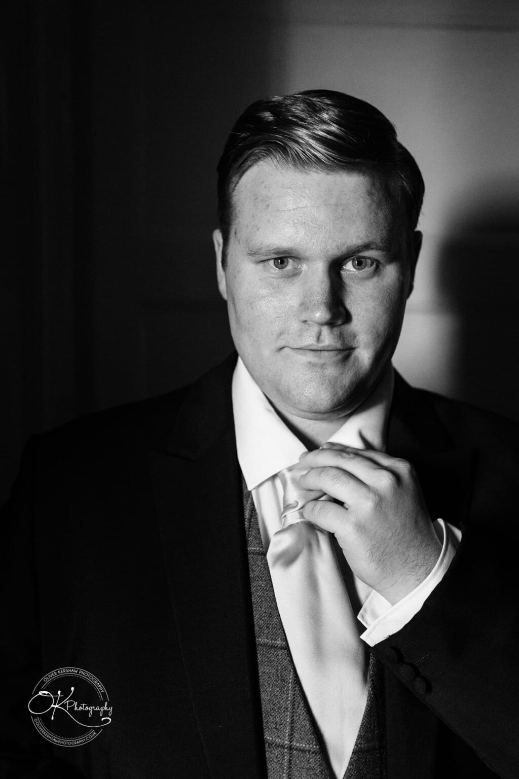 Prestwold Hall Wedding Photography Black and white portrait of a man in formal attire adjusting his tie, with a spotlight highlighting his face against a dark background.