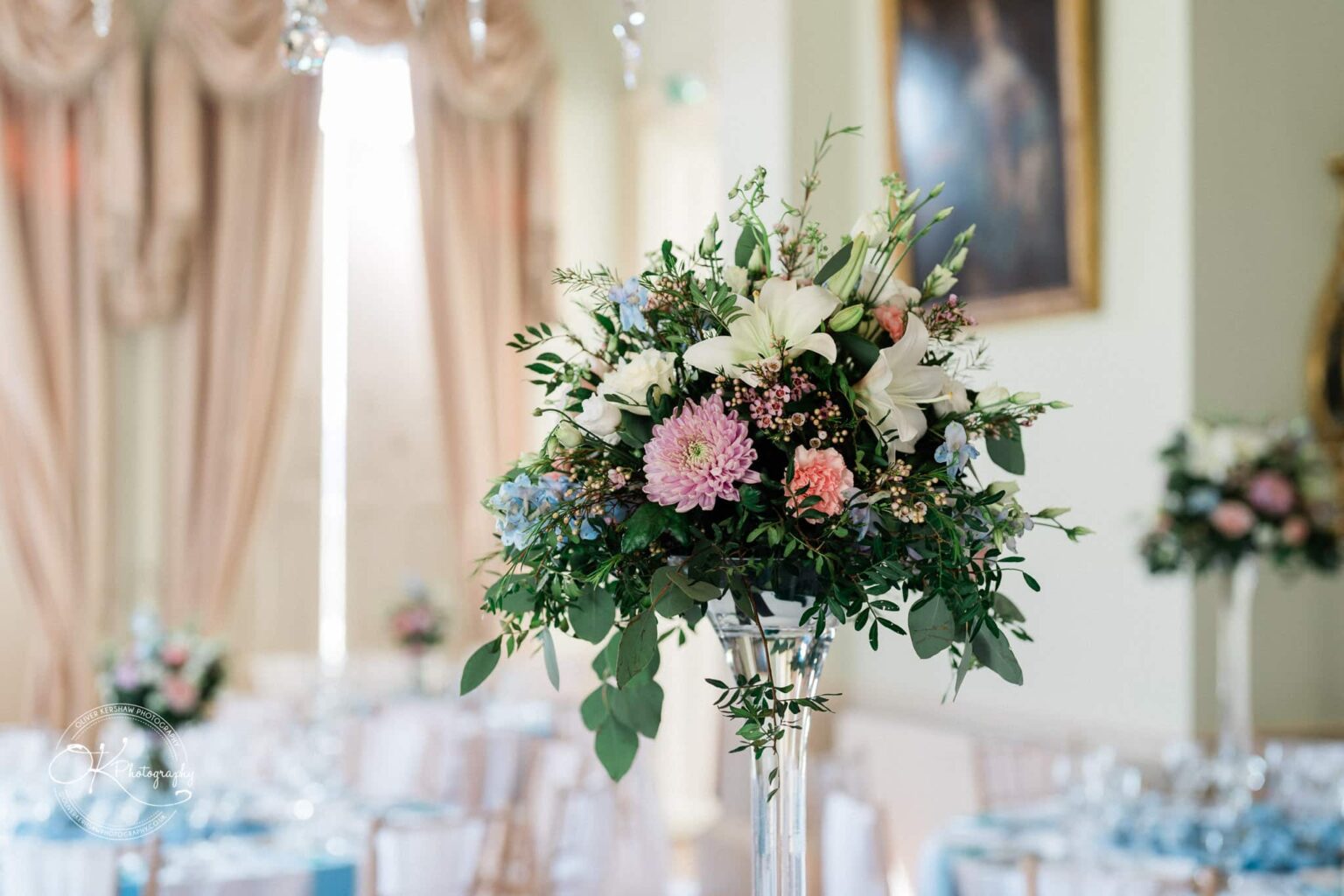 Prestwold Hall Wedding Photography Elegant floral arrangement centerpiece with pink, white, and blue flowers in a light-filled, luxurious room with draped curtains.