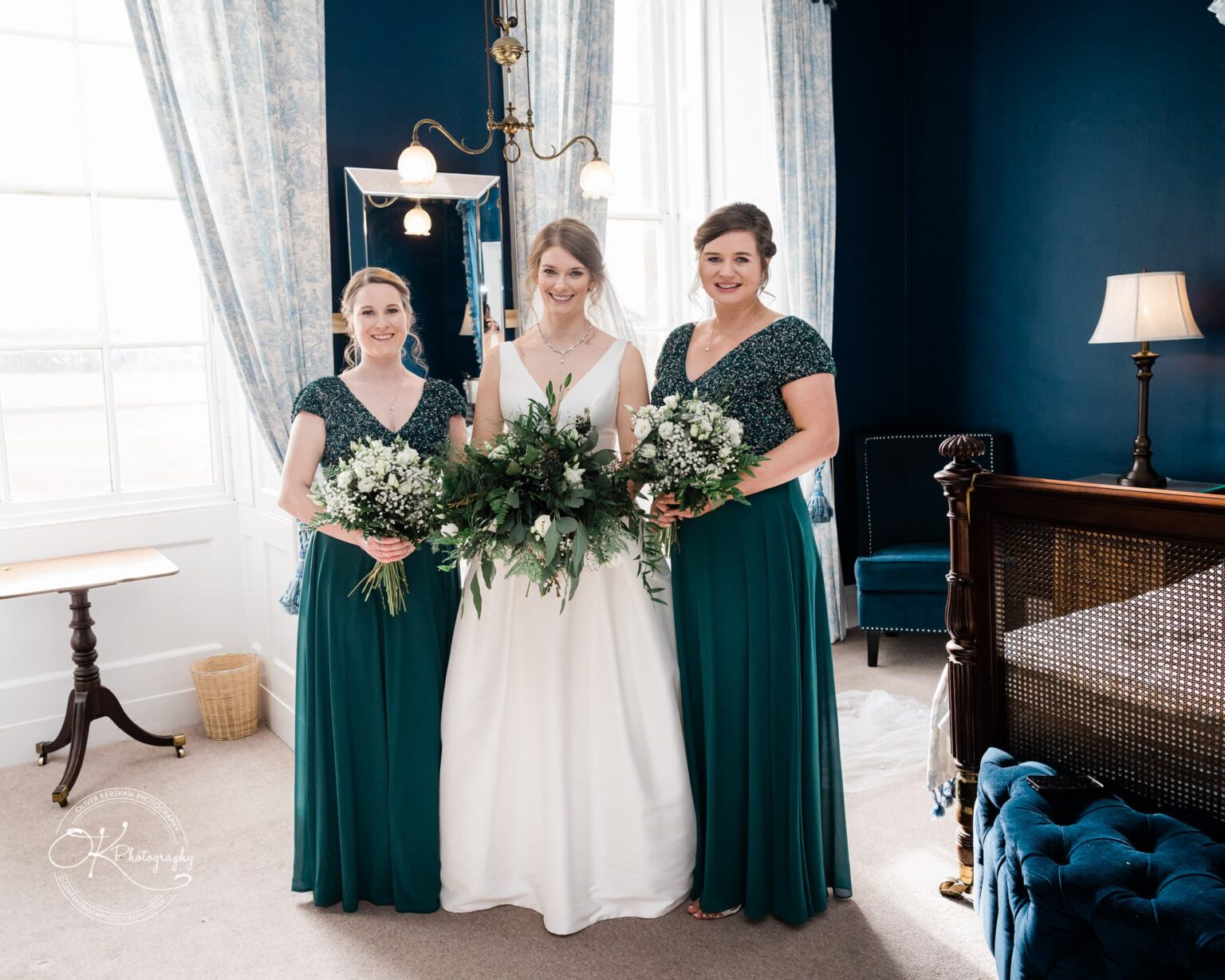 Three women in bridal attire posing in an elegant, sunlit room with blue walls and floral curtains.