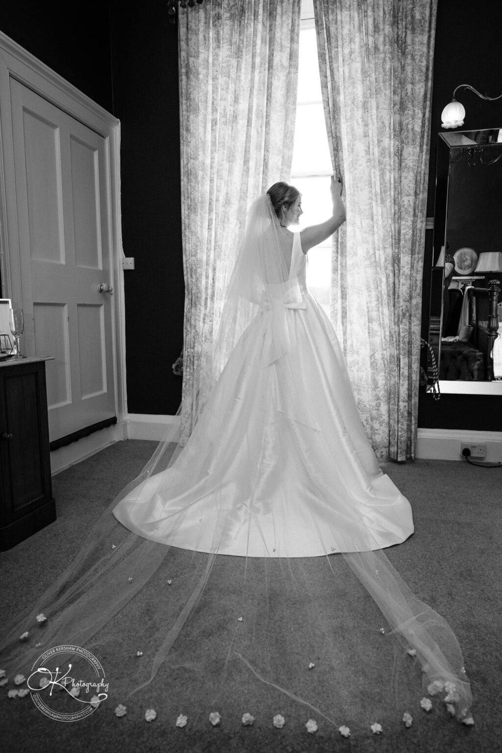 A bride in a wedding gown with a long veil stands by a window, holding back the curtain and gazing outside, in a room with elegant decor.