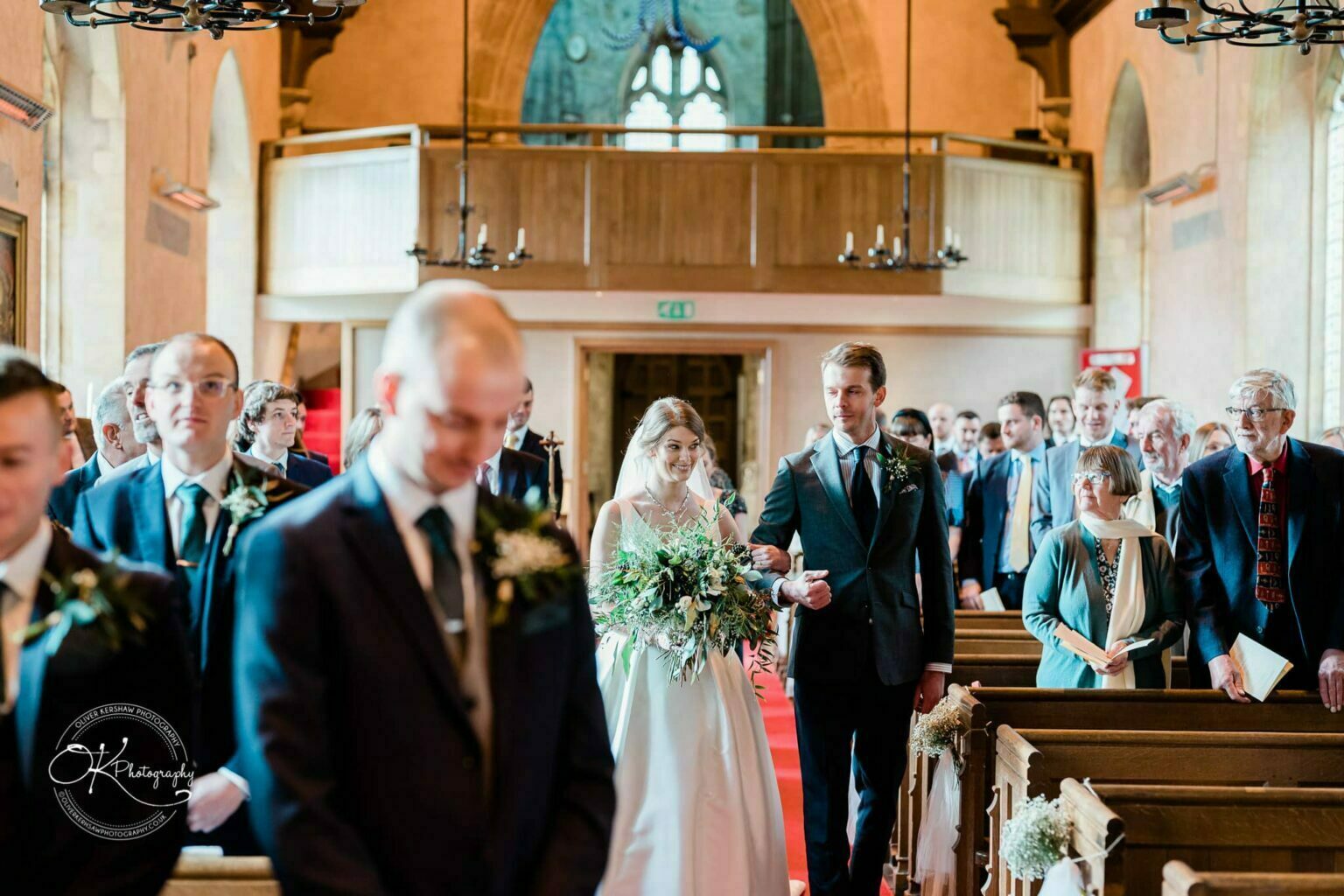 A bride walks down the aisle with a man at a wedding ceremony, while attendees watch from wooden pews inside a church.