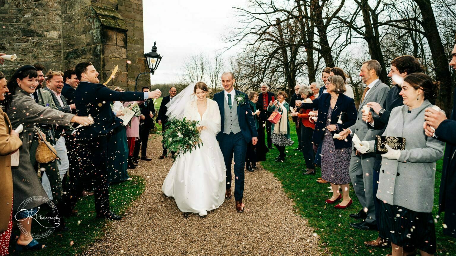 A bride and groom walking down a gravel path surrounded by guests throwing confetti.