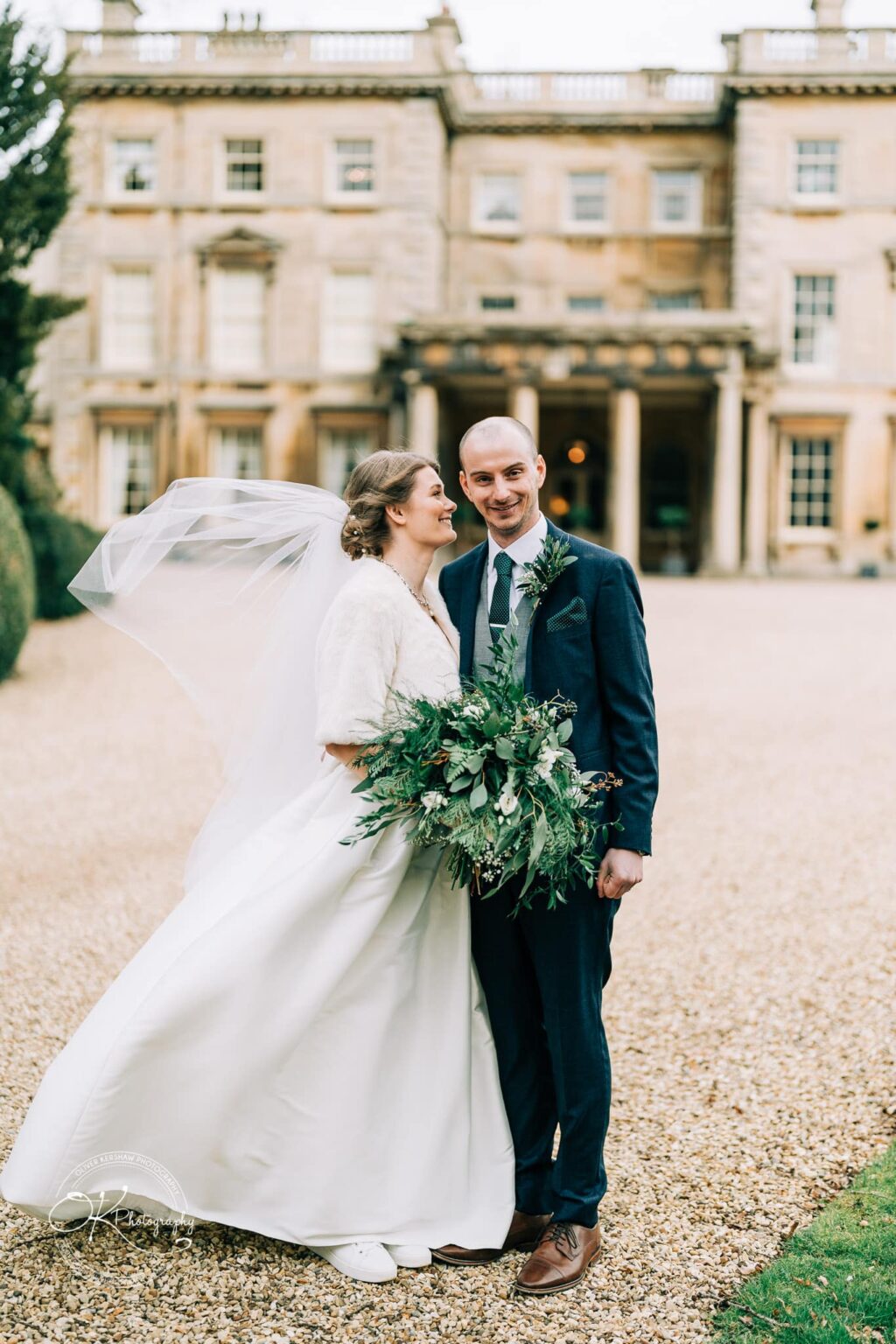 Bride and groom standing outside Prestwold Hall, with the bride holding a bouquet and her veil flowing in the wind.