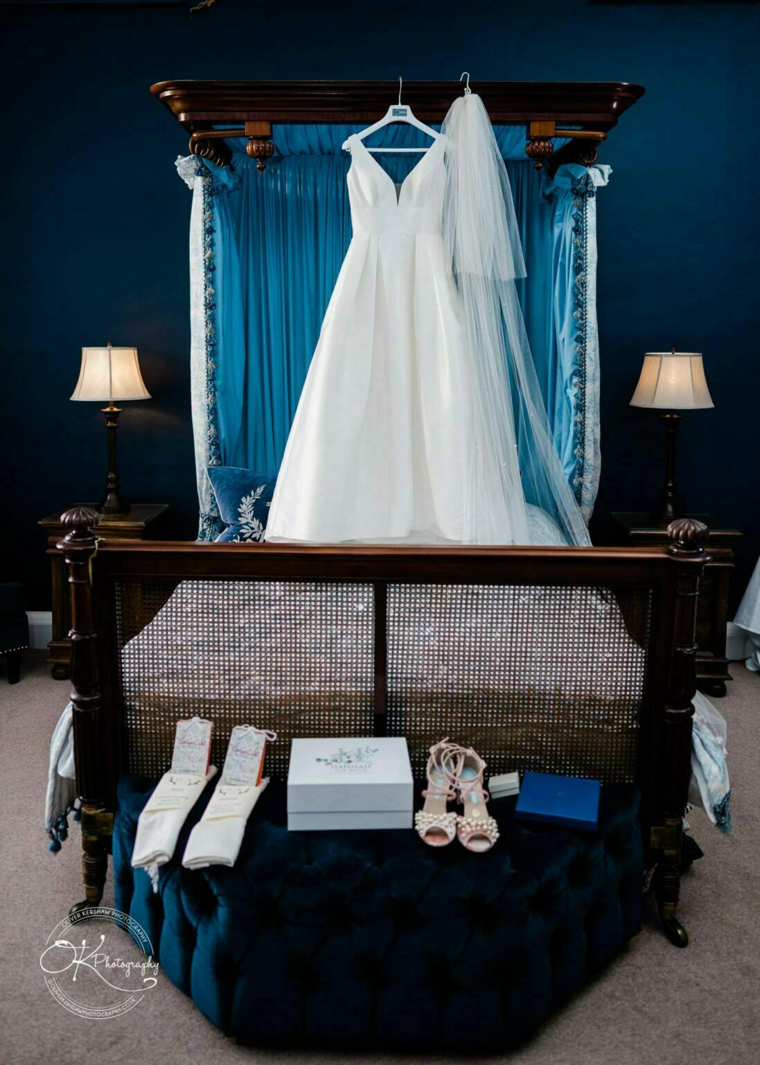 A white wedding dress with a v-neck hanging on a four-poster bed with blue drapes, accompanied by a veil. Below the bed, there are shoes, a box, and other wedding accessories, with two lamps on either side.