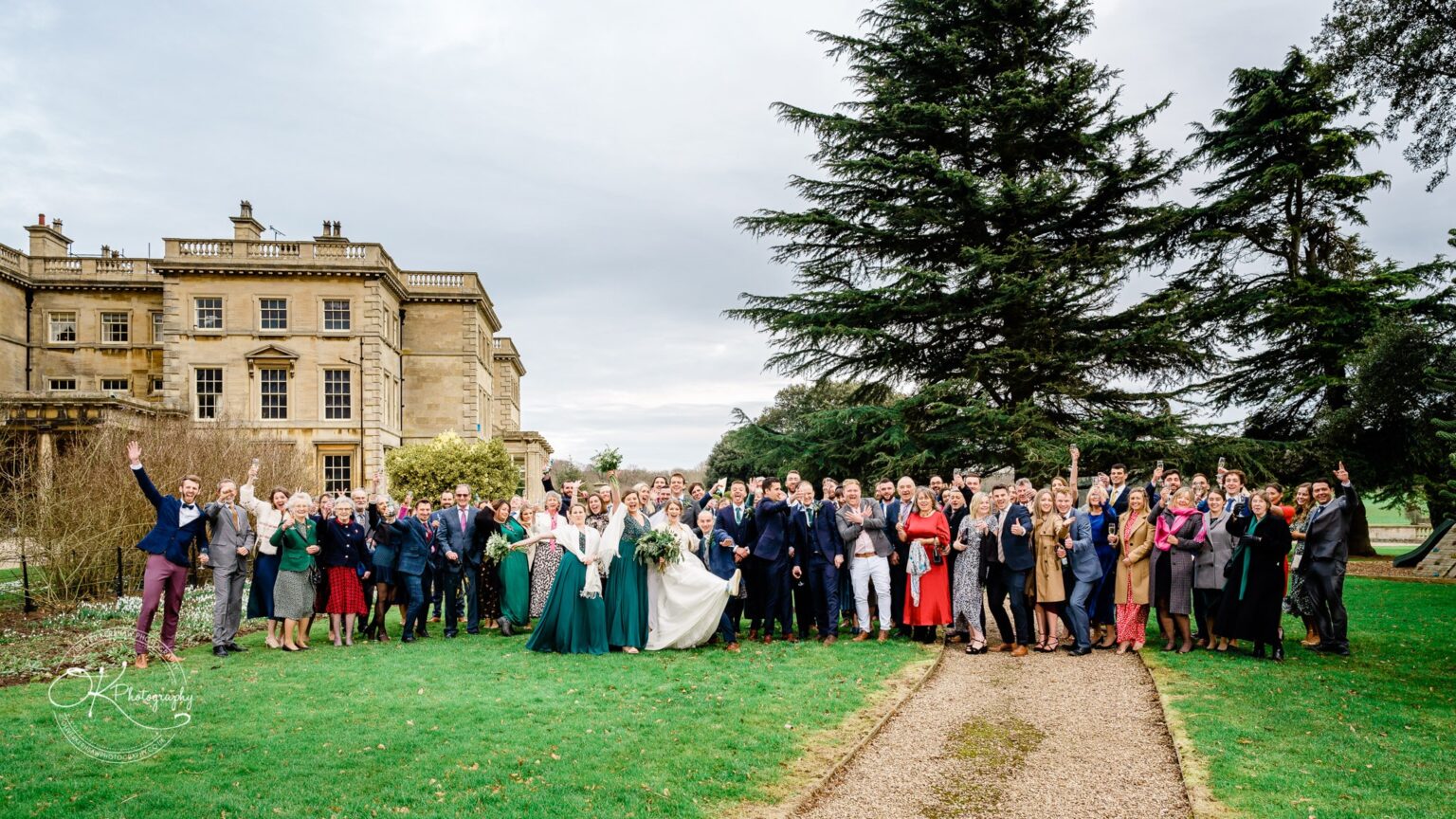 Large group of people in formal attire posing on a lawn in front of Prestwold Hall, with a large tree on the right.