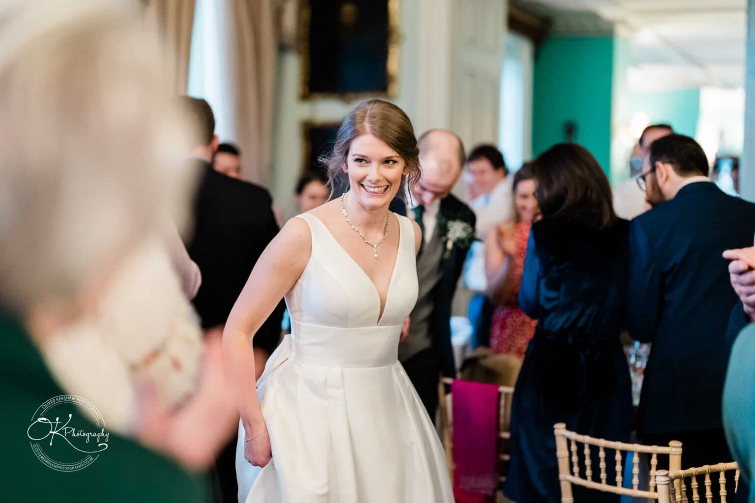 A bride in a white wedding dress is smiling as she walks past standing guests at a reception.