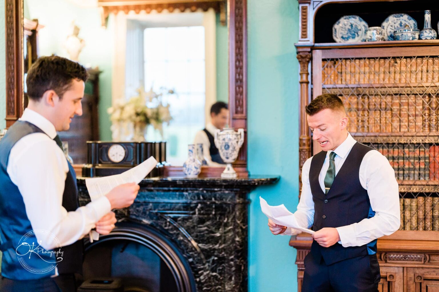 Two men in formal attire reading papers in a well-decorated room with a bookcase and ornate mantel, possibly at Prestwold Hall.