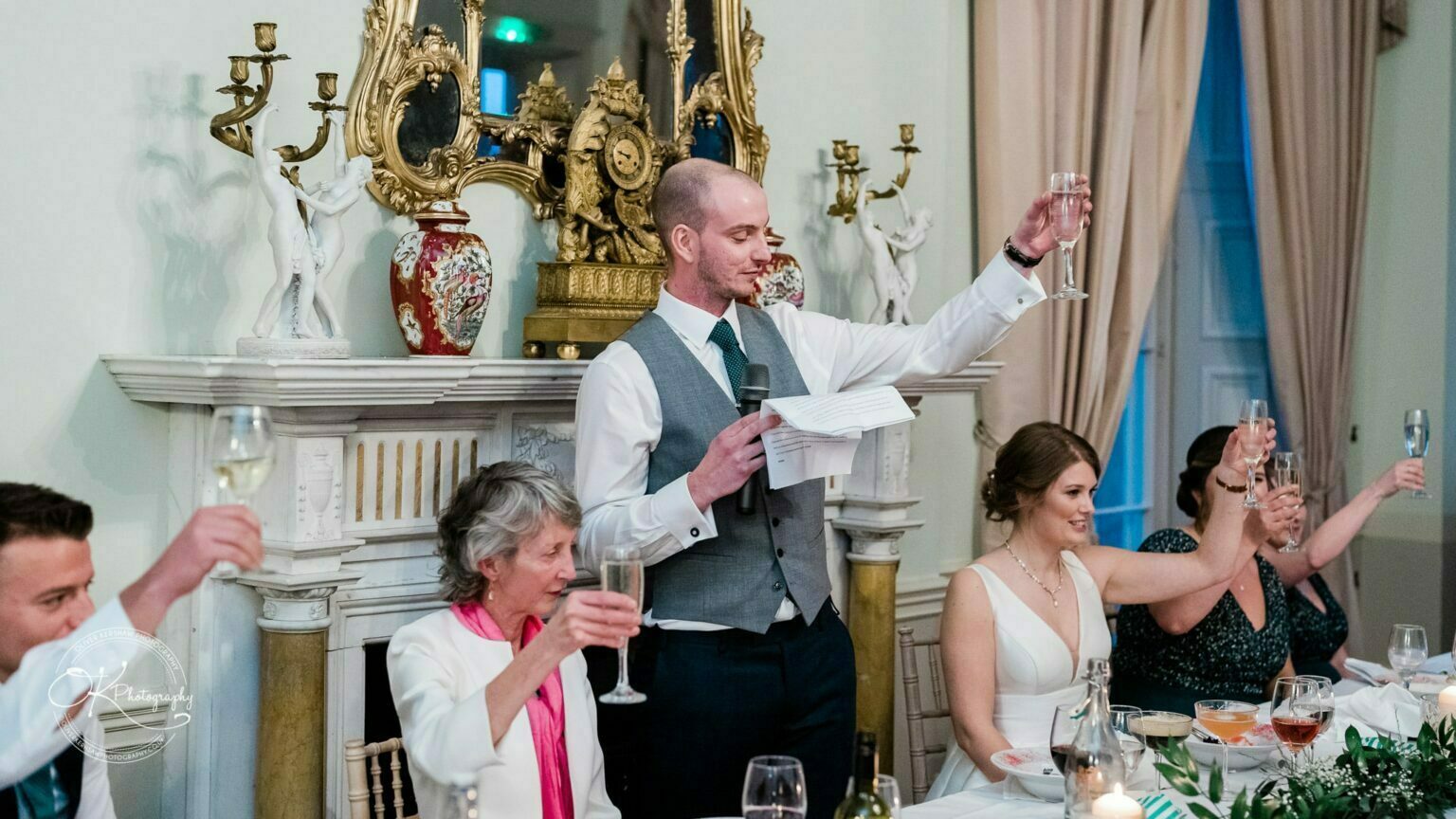 A wedding reception at Prestwold Hall with a man in a grey waistcoat giving a speech while raising a glass, and guests toasting with champagne.