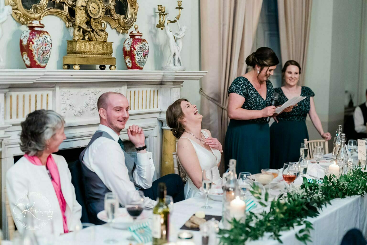 A wedding speech taking place at Prestwold Hall, with a bride laughing, a groom smiling, and other people seated at a decorated table.