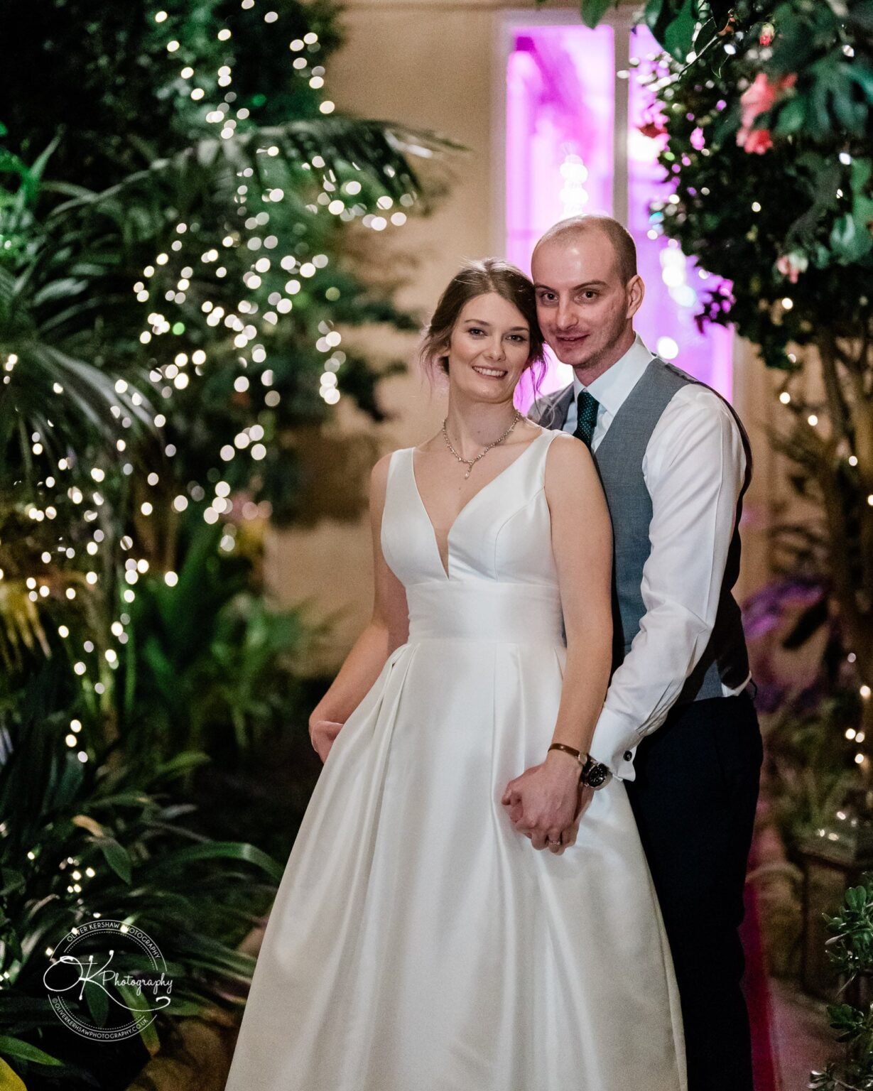 A bride and groom smiling and posing together indoors, surrounded by plants and string lights.