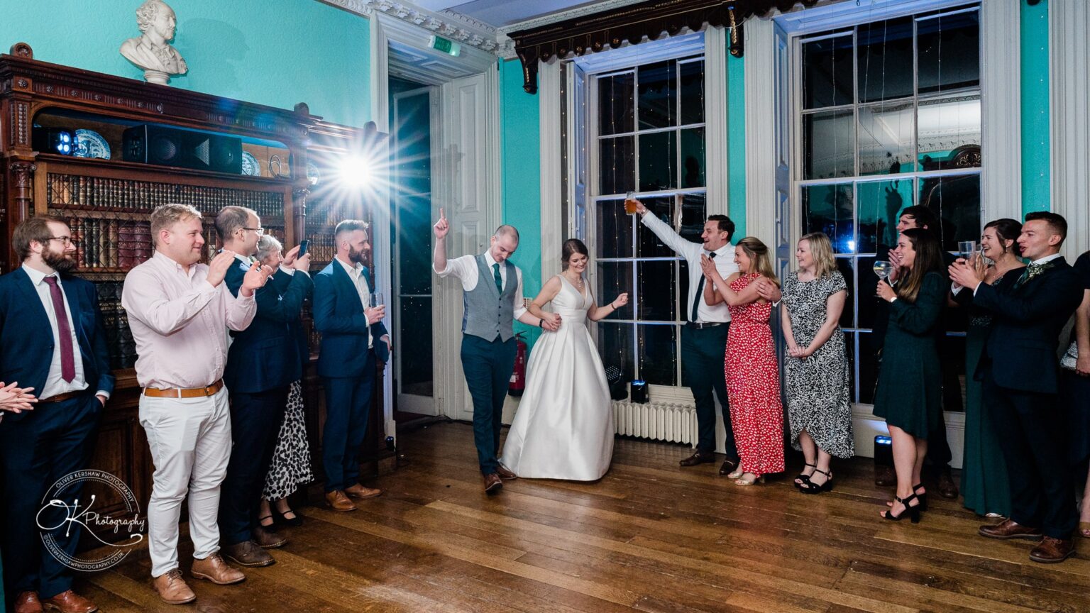 A wedding couple is dancing in a room at Prestwold Hall, surrounded by cheering guests.