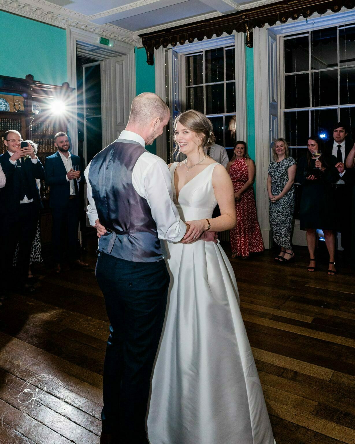 A bride and groom dance together in a room with wooden floors, while guests watch and take photos.