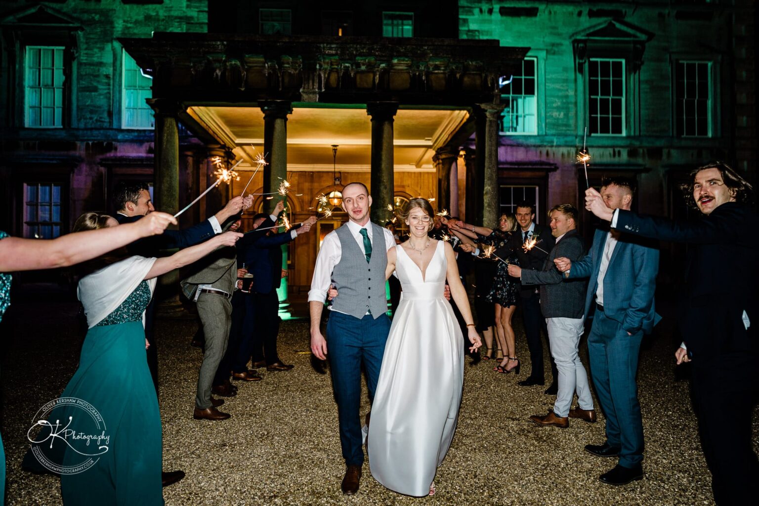 A bride and groom walking through an arch of guests holding sparklers at a nighttime wedding celebration outside Prestwold Hall.