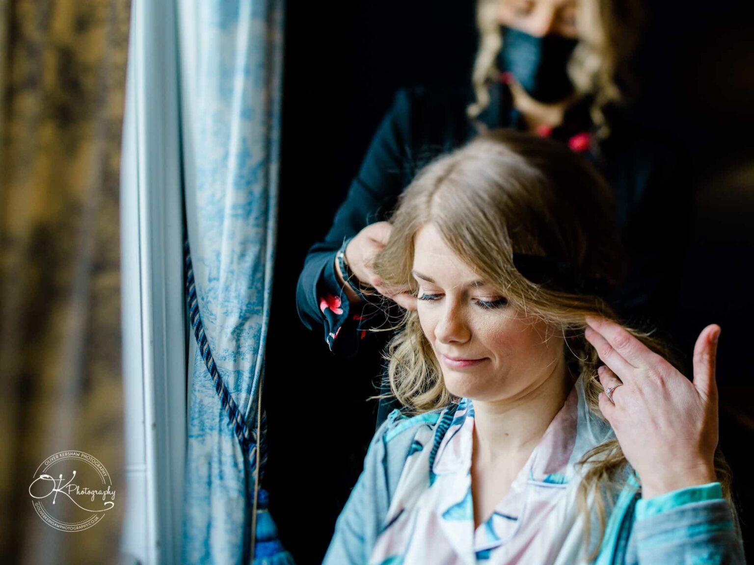Woman having her hair styled by another woman near a window with blue curtains.