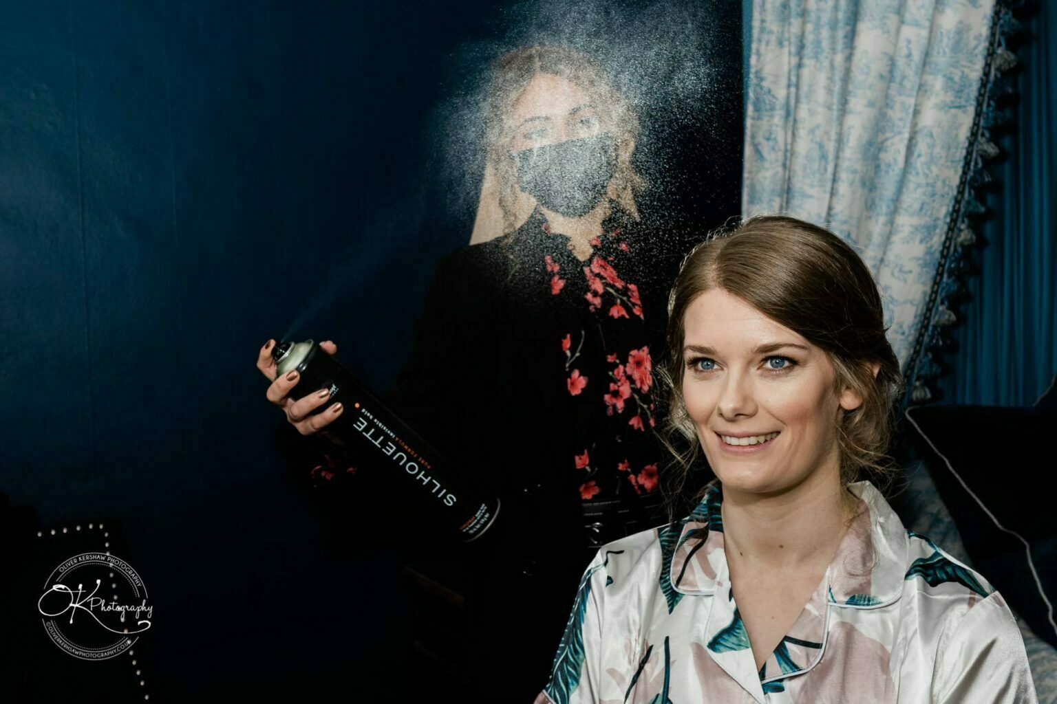 A woman in a floral dress shirt is having her hair sprayed by another woman wearing a black face mask and holding a large can of hairspray, with a dark blue background and light blue curtains.