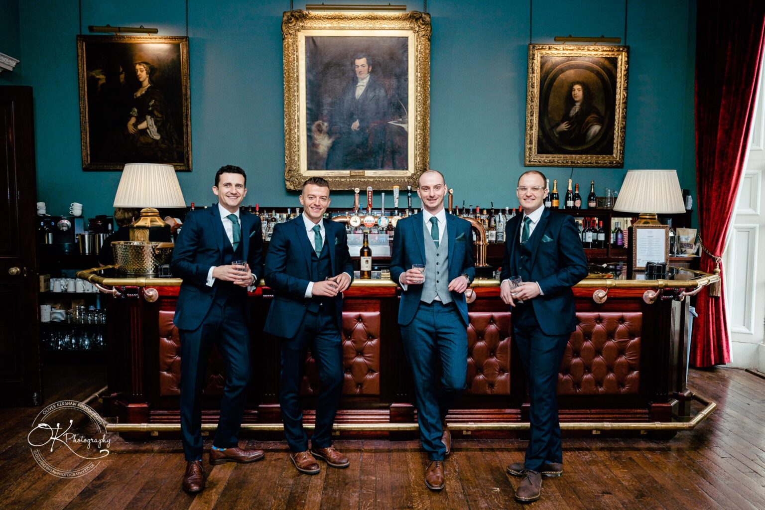 Four men in suits standing at a bar inside a room with blue walls, wooden floor, large framed portraits, lamps, and red curtains.