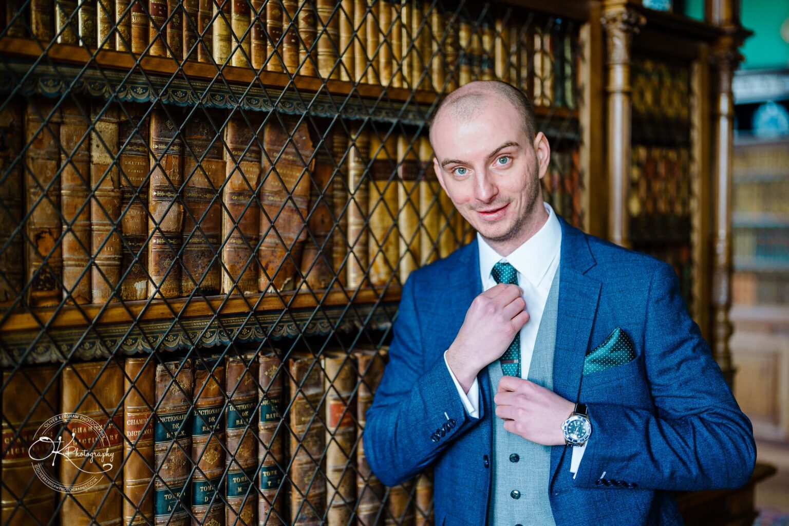 A man in a blue suit and green tie standing in front of a wooden bookshelf filled with vintage books secured behind a metal lattice at Prestwold Hall.