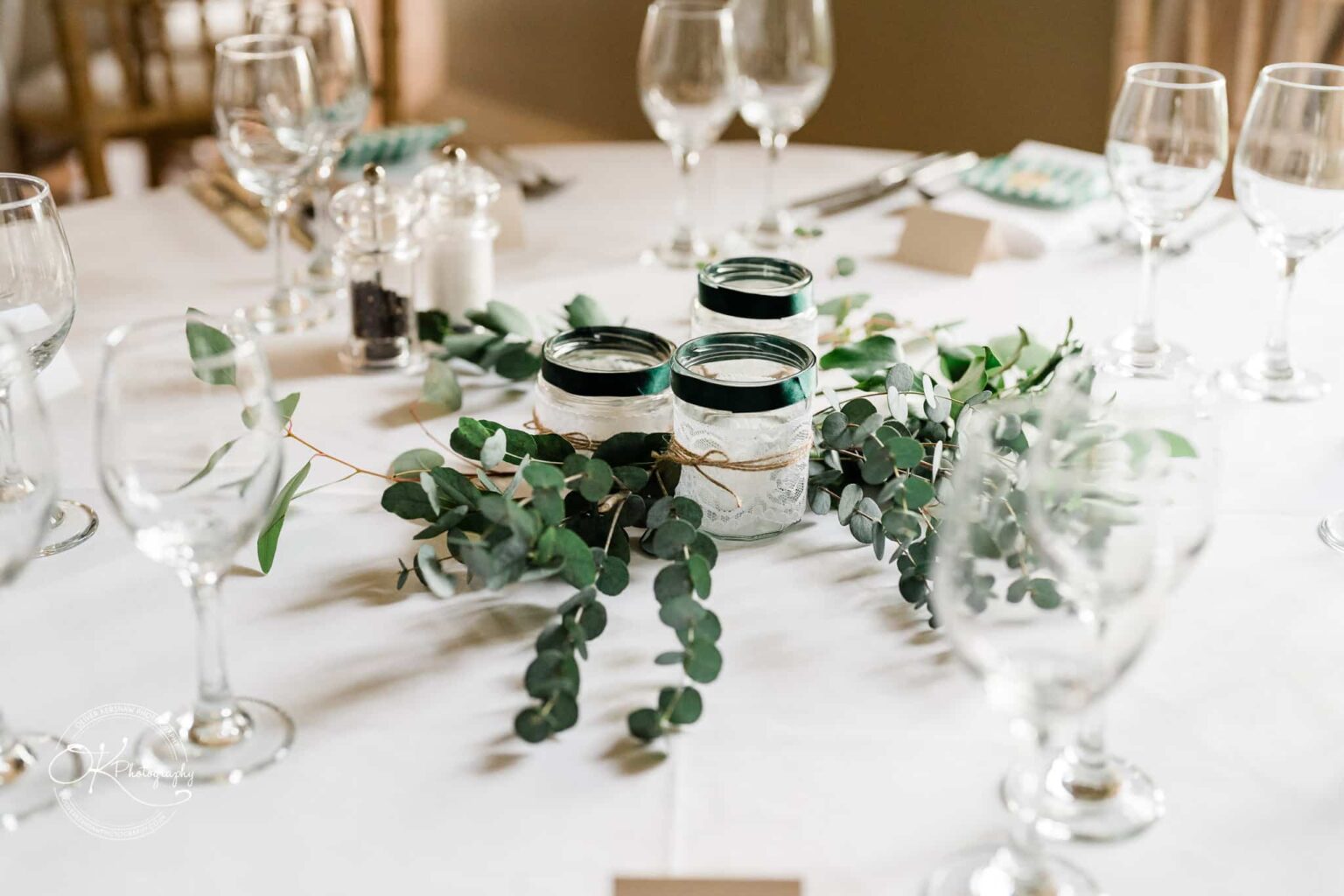 Elegant table setting with glassware, cutlery, and a centrepiece of greenery and mason jars styled with lace and twine.