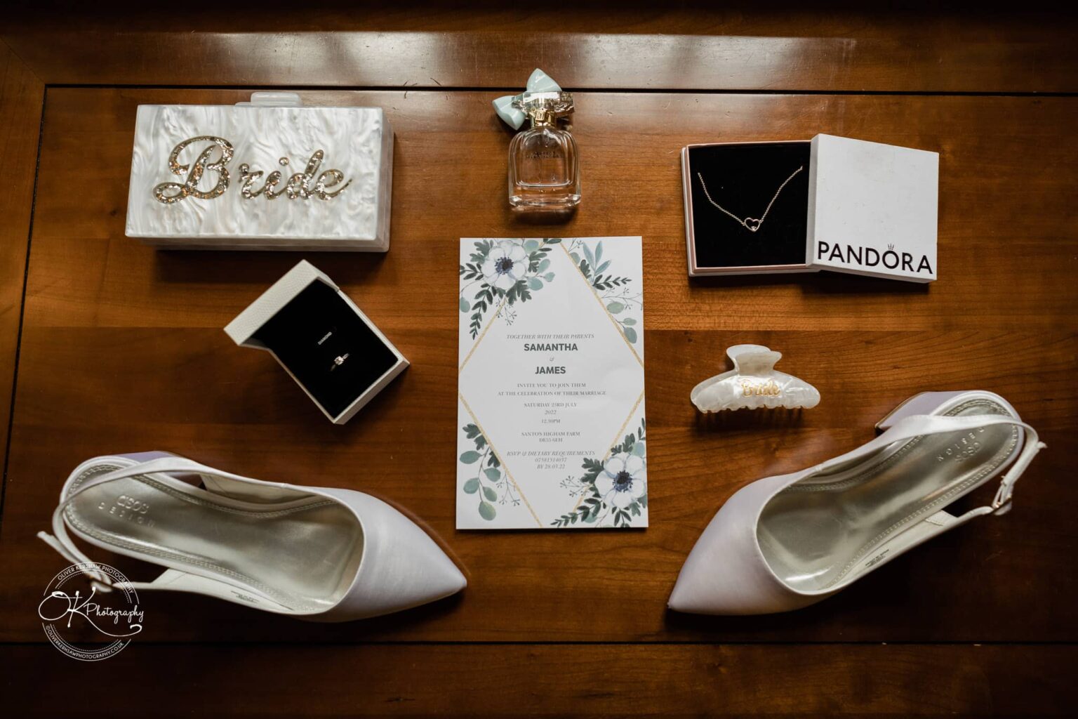 Santos Higham Farm Wedding Photography Flat lay of wedding details including a white clutch with "Bride" in rhinestones, pearl-embellished hair clip, wedding rings, Pandora necklace, perfume bottle, wedding invitation, and white bridal shoes.