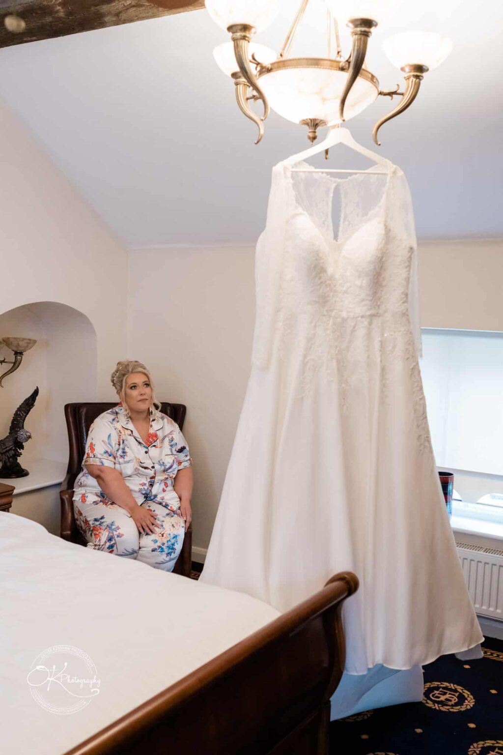 Santos Higham Farm Wedding Photography A woman in floral pyjamas sits on a chair in a bedroom, looking at a white wedding dress hanging from a chandelier.