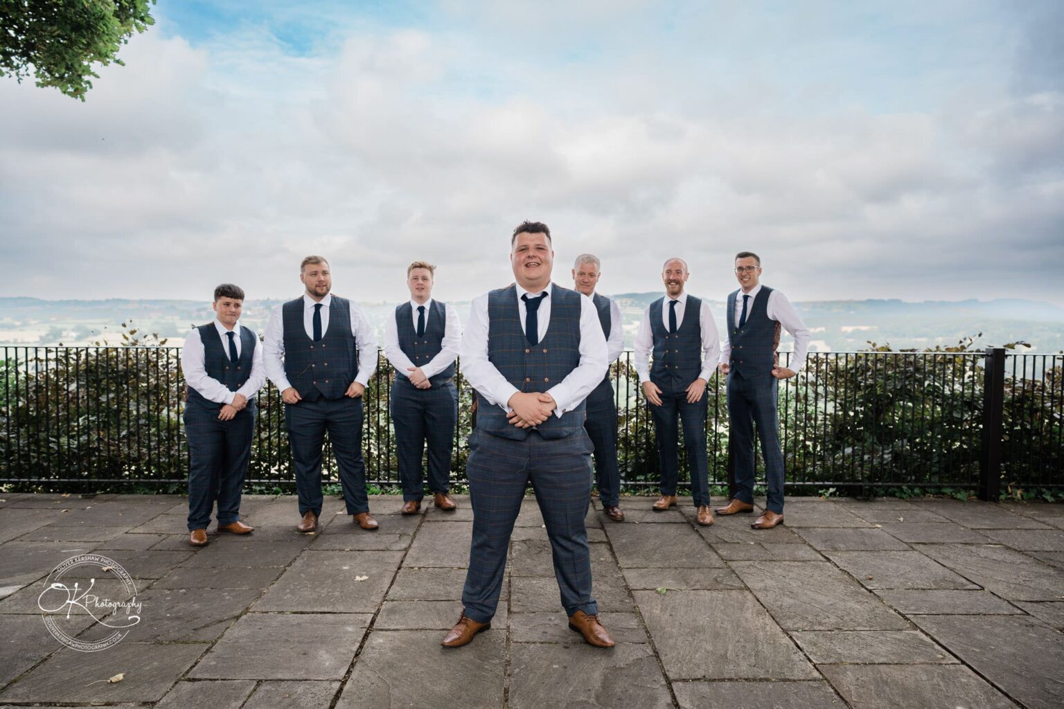 Santos Higham Farm Wedding Photography A group of seven men in matching suits and ties, standing outdoors with a scenic view and a cloudy sky in the background.