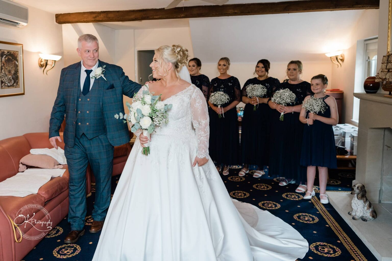 Santos Higham Farm Wedding Photography A bride in a white wedding dress holding a bouquet stands next to a man in a blue checked suit, with bridesmaids in navy blue dresses holding flowers in the background.