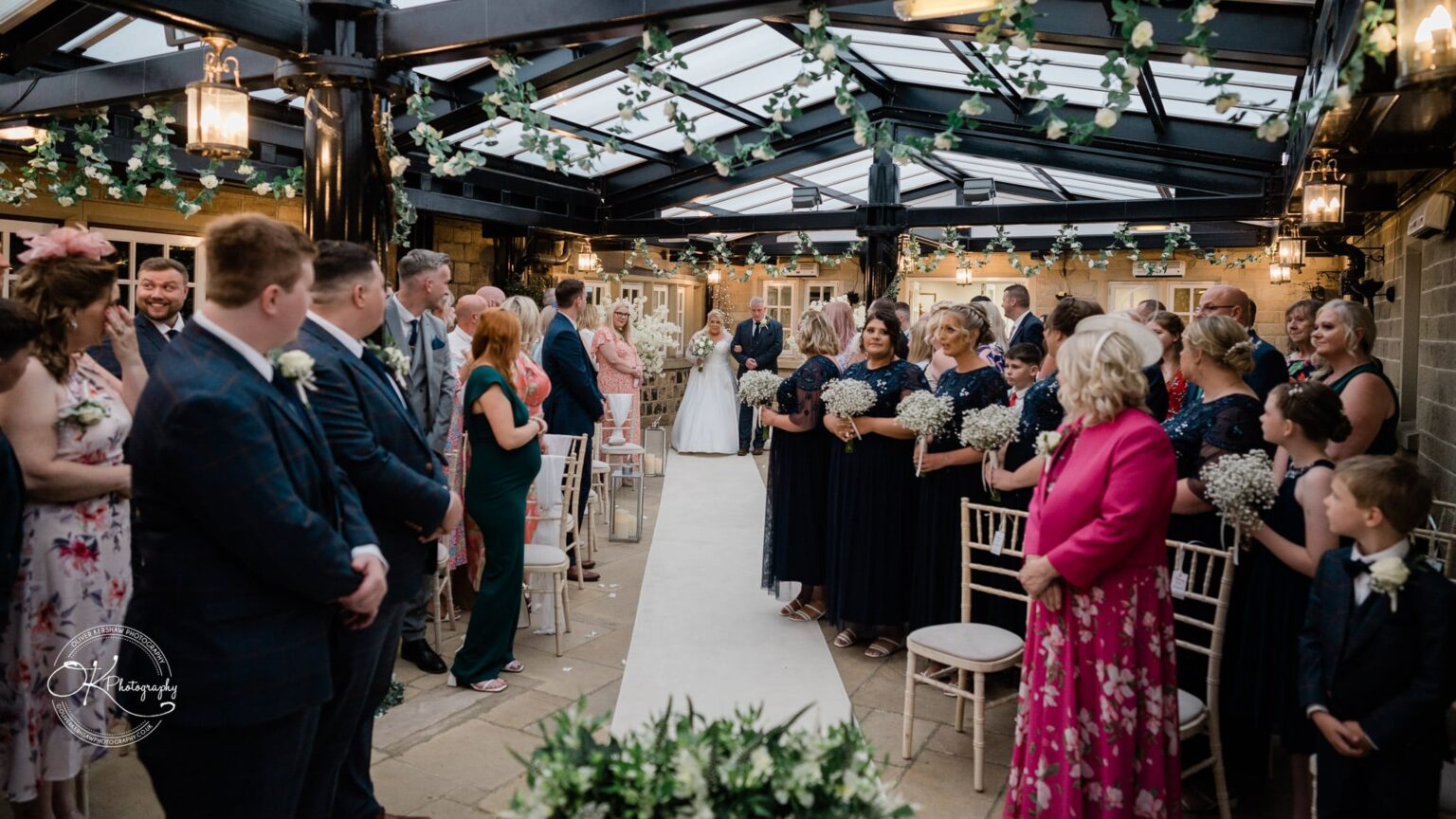Santos Higham Farm Wedding Photography Indoor wedding ceremony with guests standing on either side of an aisle adorned with greenery, as the bride walks down the aisle with her father.