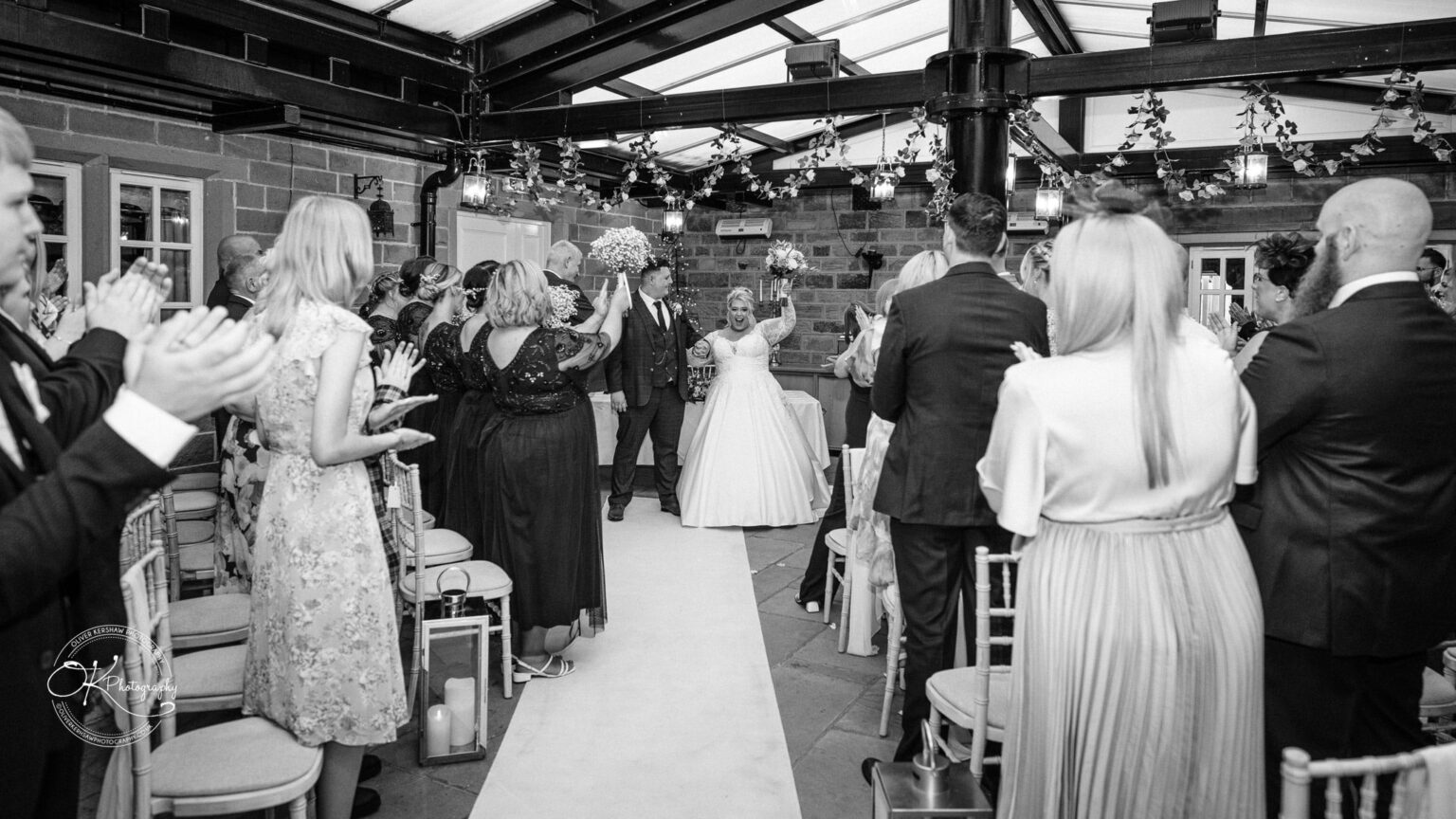 Santos Higham Farm Wedding Photography A bride and groom stand at the end of an aisle in a decorated venue, surrounded by applauding guests.