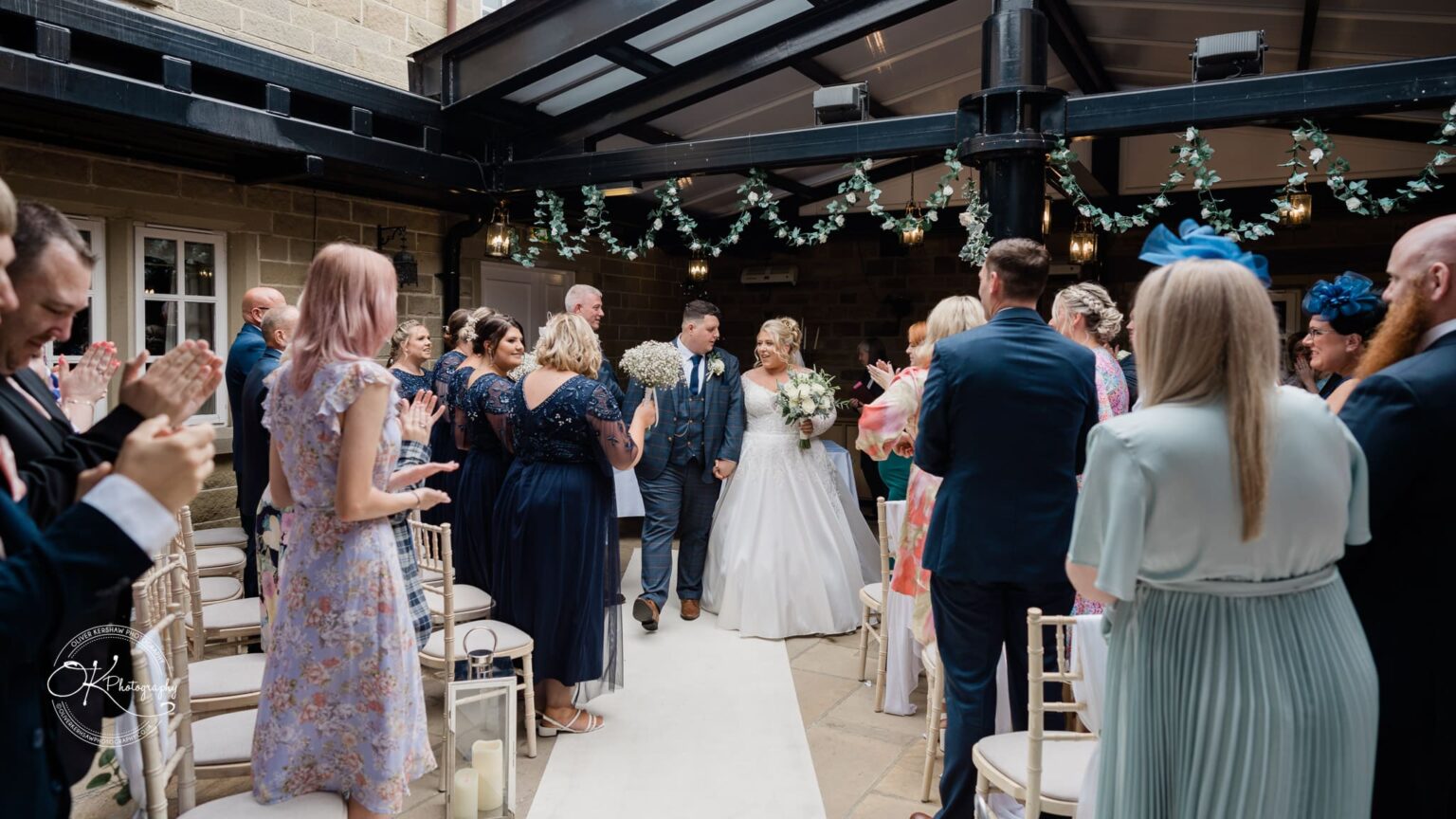 Santos Higham Farm Wedding Photography Bride and groom walking down the aisle at their wedding ceremony, surrounded by applauding guests, with bridesmaids in navy dresses holding bouquets.