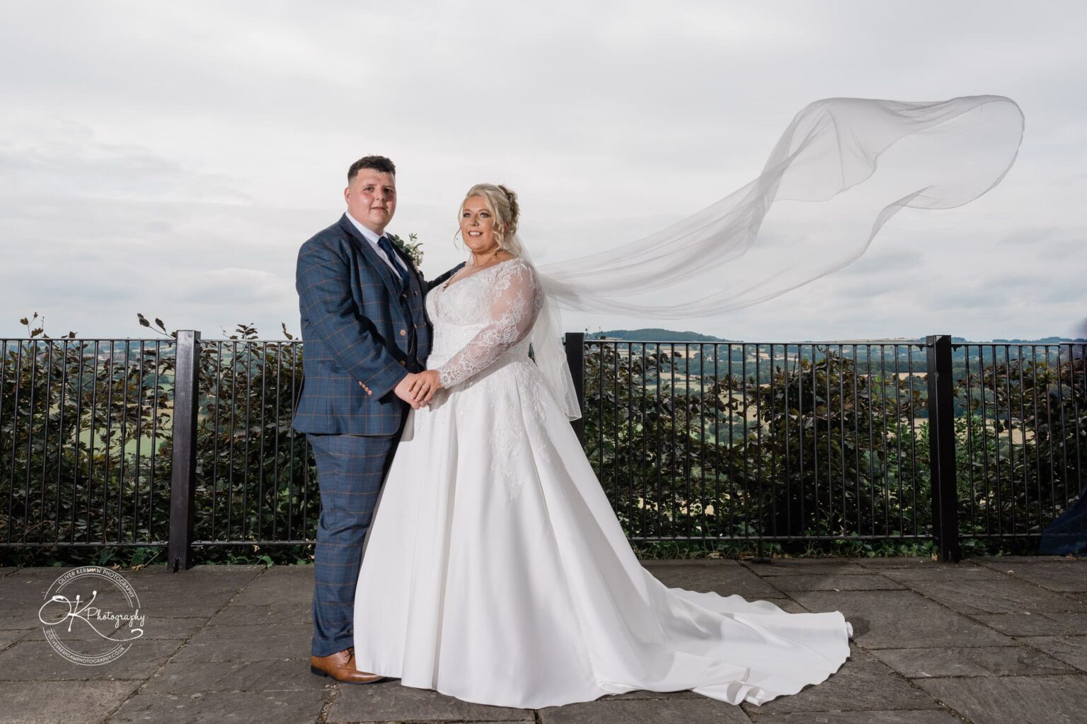 Santos Higham Farm Wedding Photography Bride and groom standing outdoors, the bride's dress train and veil flowing elegantly, with a scenic background of greenery and hills.