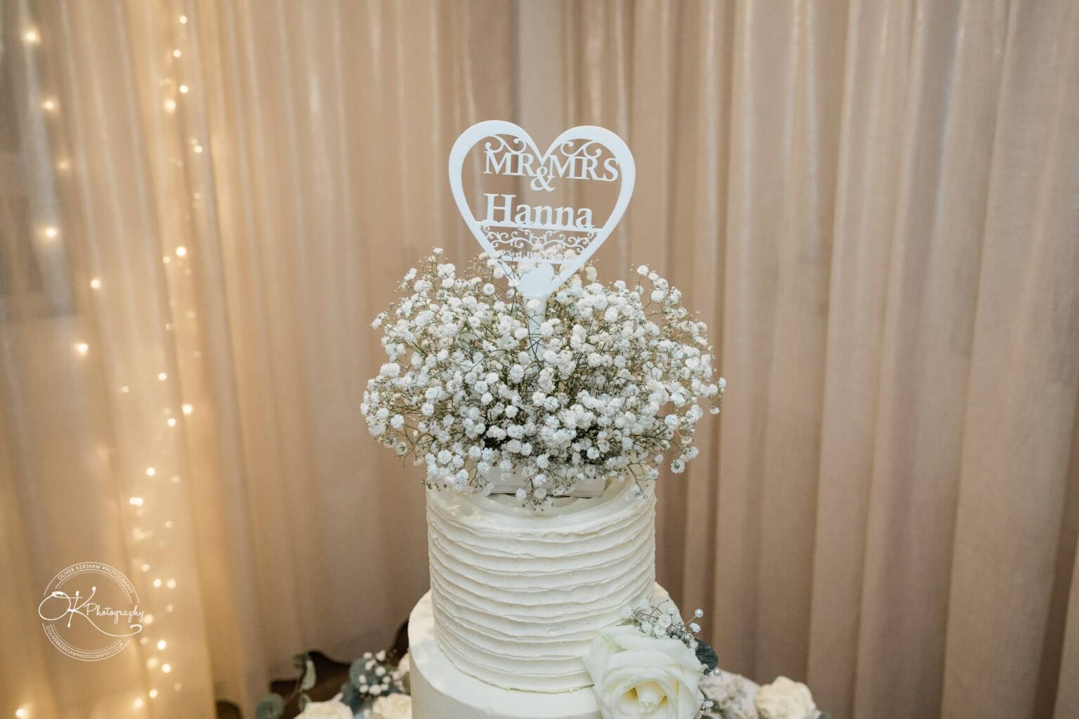 Santos Higham Farm Wedding Photography A white wedding cake with a "Mr. & Mrs. Hanna" heart-shaped topper, decorated with baby's breath flowers and a white rose at the base, set against a backdrop of sheer curtains with fairy lights.