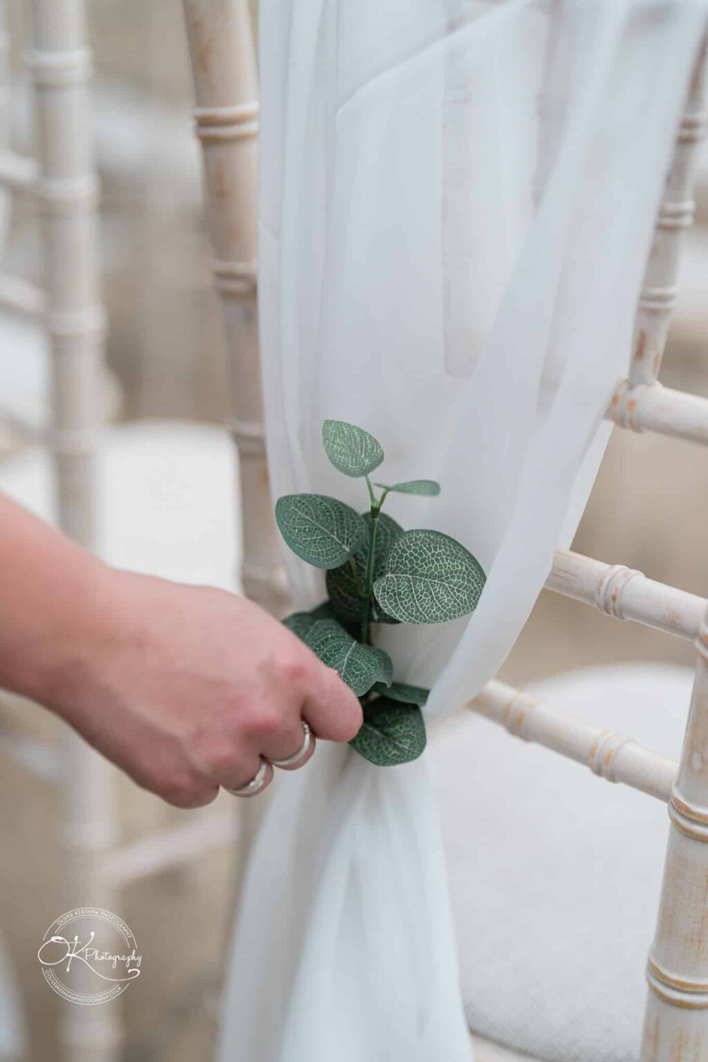 Santos Higham Farm Wedding Photography A hand adjusting a small sprig of green leaves tied with white fabric on a bamboo chair.