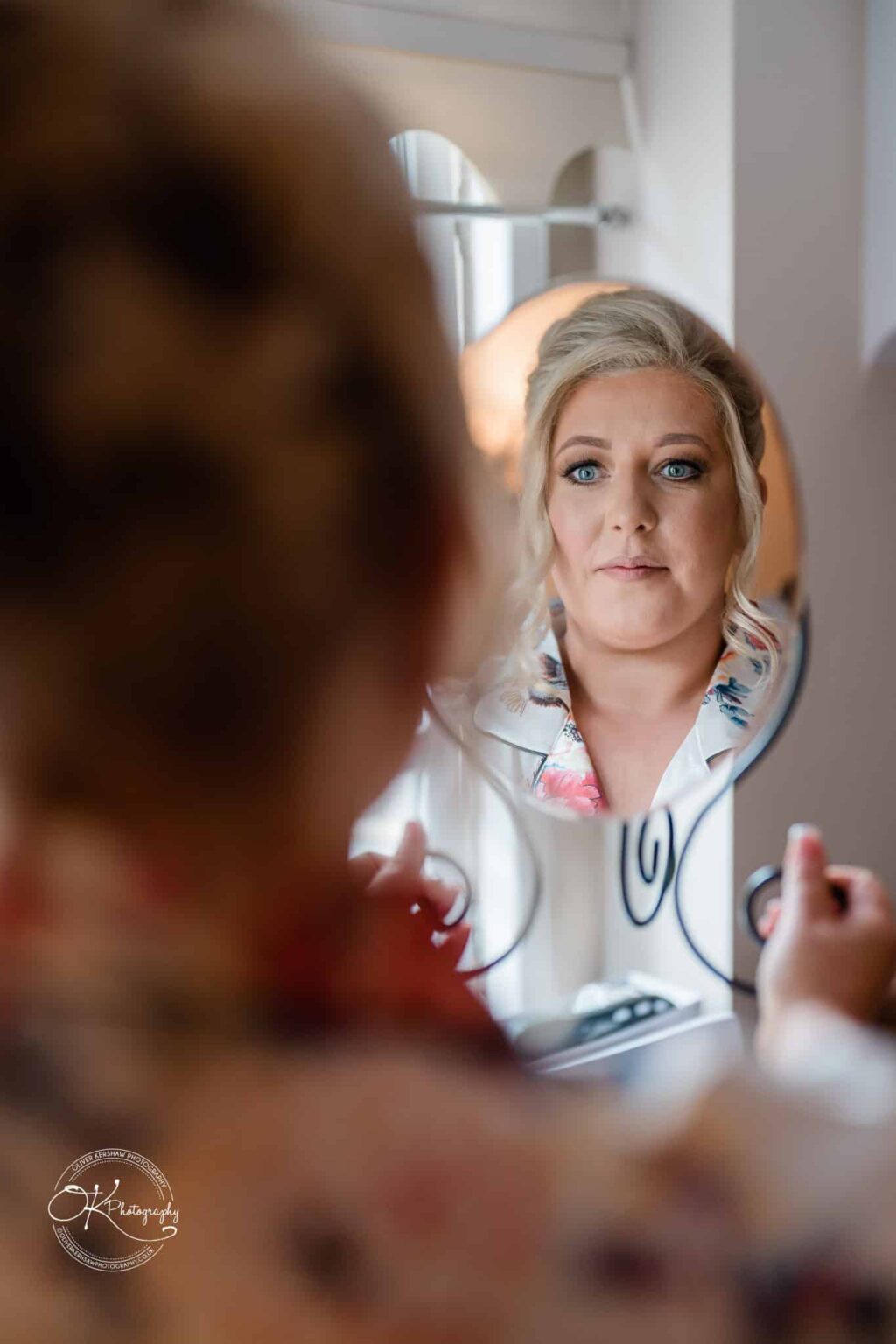 Santos Higham Farm Wedding Photography Woman looking at herself in a round mirror while holding makeup, seen from over her shoulder.