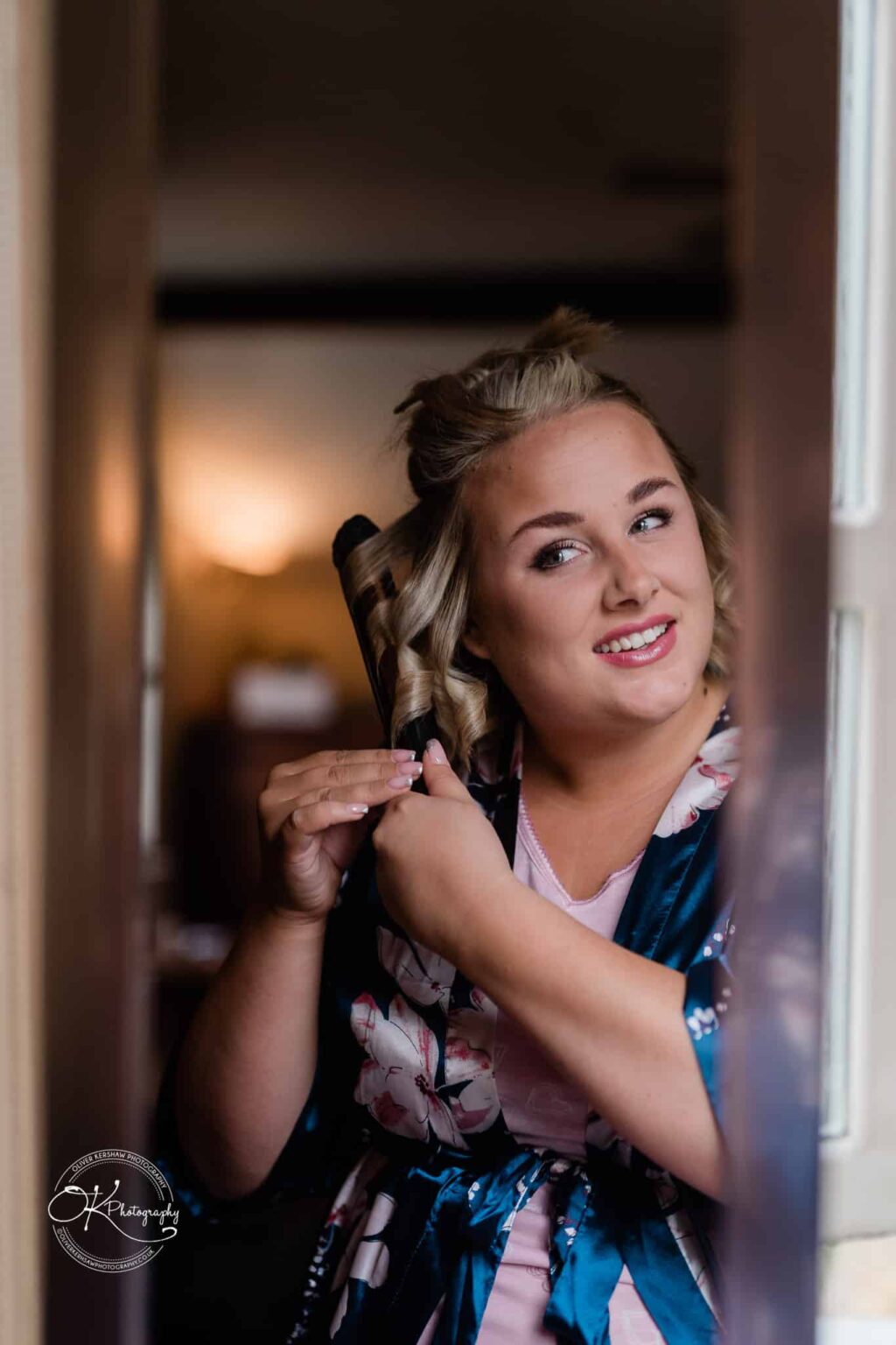 Santos Higham Farm Wedding Photography A woman curling her hair while looking in a mirror. She is smiling and wearing a floral robe.