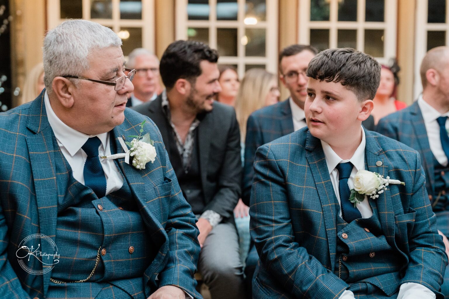 Santos Higham Farm Wedding Photography An older man and a boy, both wearing blue checked suits with white rose boutonnieres, are sitting and looking at each other. Several people are seated in the background.