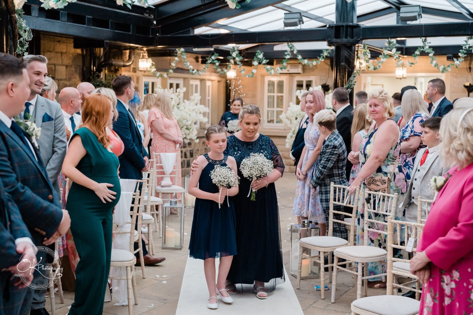 Santos Higham Farm Wedding Photography Wedding ceremony with a group of guests standing and watching a woman and a girl holding flower bouquets walking down an aisle lined with white chairs.